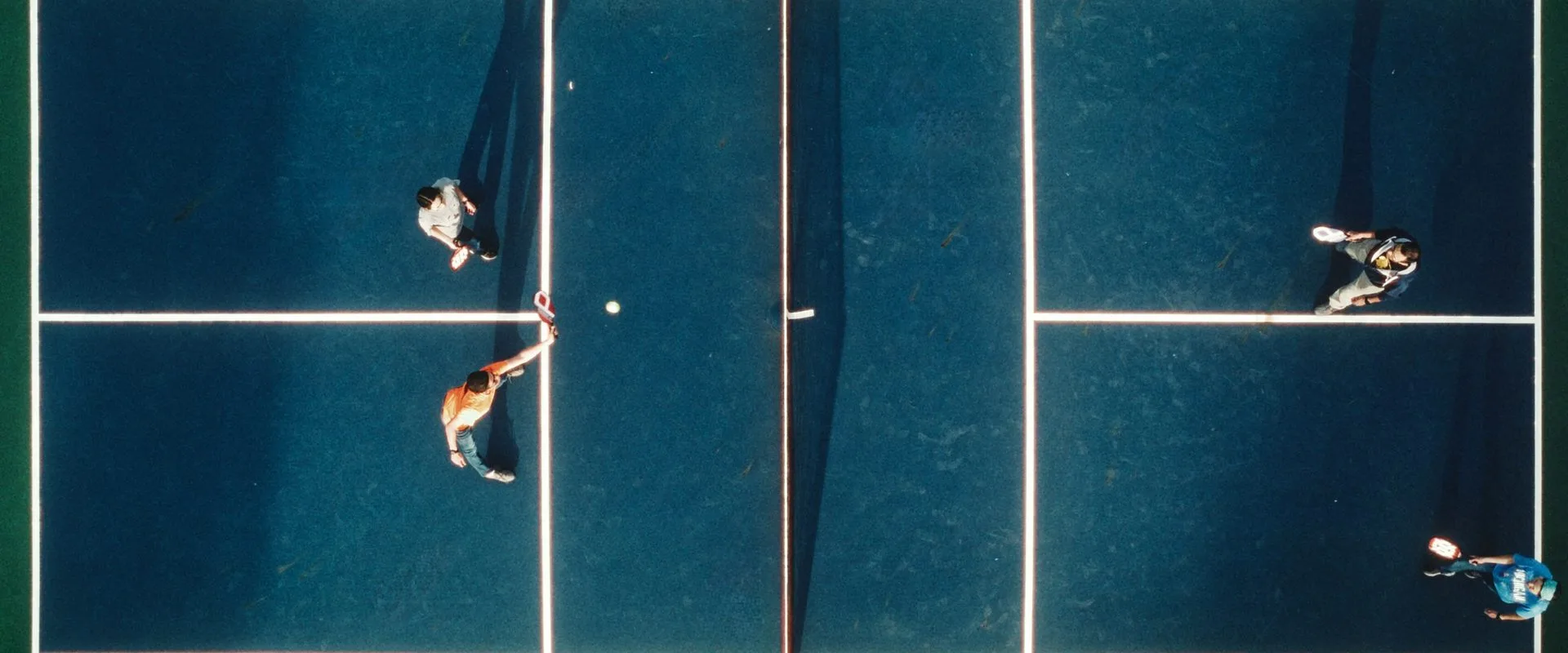 Aerial view of four players in a doubles game on an outdoor pickleball court, with one net across the middle and white court lines